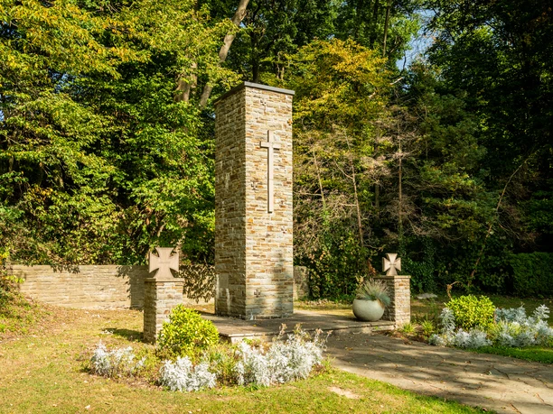 Streifzug Kräuterweg Gedenkstätte aus Stein mit Kreuz, umgeben von Bäumen und Pflanzen in Neunkirchen-Seelscheid.