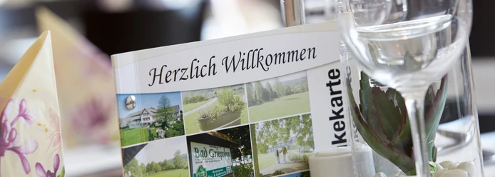 Hotel-Bad-Griepshop Menükarte mit der Aufschrift "Herzlich Willkommen" in einem Hotelrestaurant, neben einem Weißweinglas.