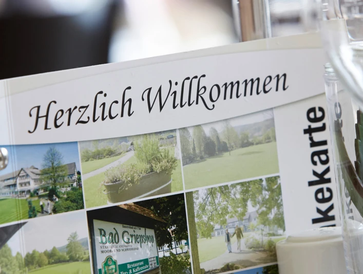 Hotel-Bad-Griepshop Menükarte mit der Aufschrift "Herzlich Willkommen" in einem Hotelrestaurant, neben einem Weißweinglas.