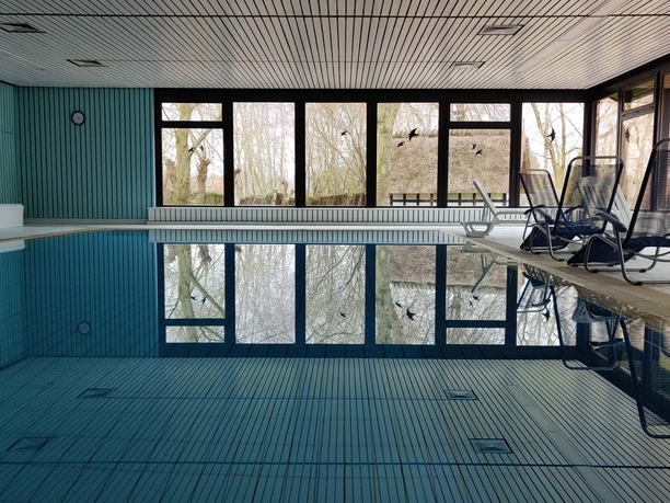Kurhaus Pivittskrug Innenansicht eines Hallenbads mit großen Fenstern, Liegestühlen und Spiegelung im blauen Wasser.