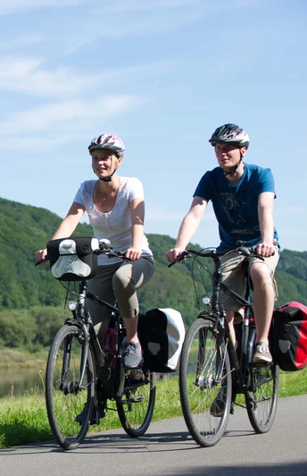 Mit dem Fahrrad entlag der Weser unterwegs Zwei Personen radeln auf einem Fahrradweg entlang der Weser, umgeben von üppigen, grünen Wäldern.