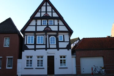 Hotel Ambiente Fachwerkhaus mit weißer Fassade und dunklem Holzgebälk, umgeben von Ziegelbauten, vor blauem Himmel.