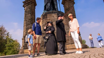 Kaiser-Wilhelm-Denkmal Historische Führung.jpg Personen jeden Alters lauschen einem historischen Führer am Kaiser-Wilhelm-Denkmal auf Kopfsteinpflaster.