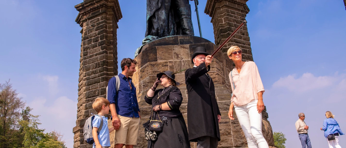 Kaiser-Wilhelm-Denkmal Historische Führung.jpg Personen jeden Alters lauschen einem historischen Führer am Kaiser-Wilhelm-Denkmal auf Kopfsteinpflaster.