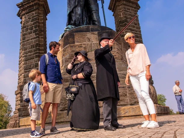 Kaiser-Wilhelm-Denkmal Historische Führung.jpg Personen jeden Alters lauschen einem historischen Führer am Kaiser-Wilhelm-Denkmal auf Kopfsteinpflaster.