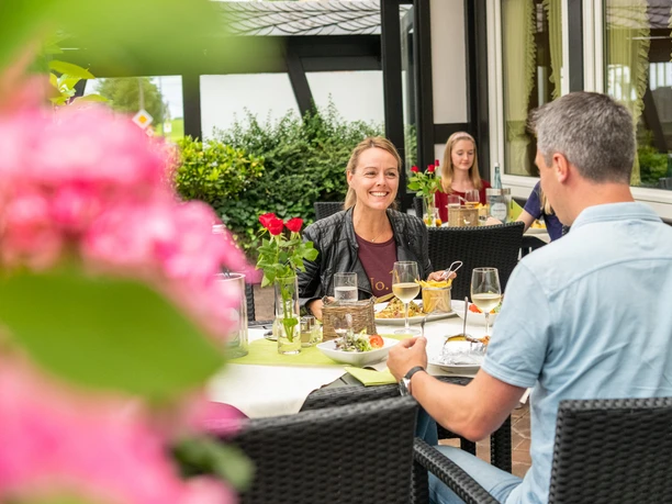 Eine Frau und ein Mann essen auf einer Terrasse, umgeben von blühenden Pflanzen.