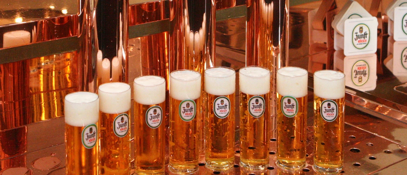Produkte aus der Region Acht Gläser Kölschbier stehen aufgereiht vor glänzenden Kupfertheken in einer Brauerei.