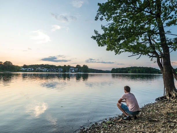 Bevertalsperre Ein Mann sitzt am Ufer eines Sees bei Sonnenuntergang, umgeben von Natur und ruhigem Wasser.