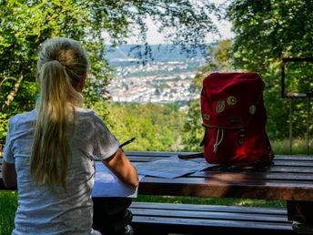 Aussicht am HAGENfenster.jpg Person mit Notizblock und rotem Rucksack, blickt von Bank auf bewaldete Landschaft und Stadt.
