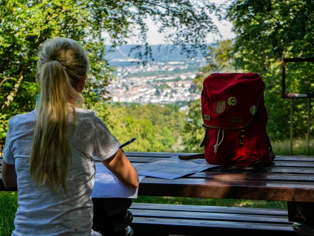 Aussicht am HAGENfenster.jpg Person mit Notizblock und rotem Rucksack, blickt von Bank auf bewaldete Landschaft und Stadt.