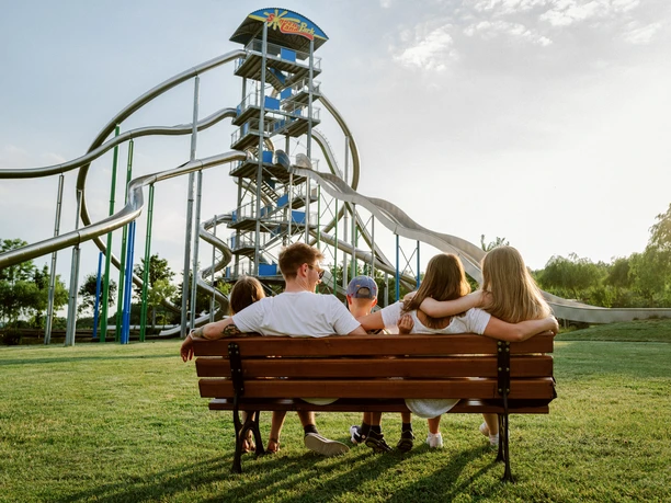 Rutschenturm Familie auf Parkbank vor hohem Rutschenturm mit mehreren Wasserrutschen im sonnigen Park.