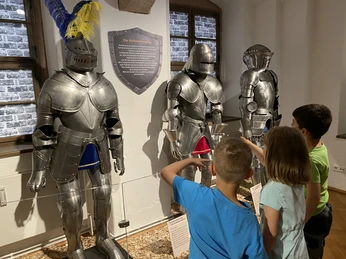 Turnierrüstungen mit der Kindern Kinder betrachten drei mittelalterliche Ritterrüstungen in einem Museum mit Infotafel.Children look at three pieces of medieval knight's armor in a museum with an information board.Děti si v muzeu s informační tabulí prohlédnou tři kusy středověké rytířské zbroje.Dzieci oglądają trzy części średniowiecznej zbroi rycerskiej w muzeum z tablicą informacyjną.Kinderen bekijken drie stukken middeleeuws ridderharnas in een museum met een informatiebord.I bambini osservano tre armature di cavalieri medievali in un museo con un pannello informativo.
