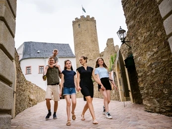 Burg Scharfenstein Eine Familie spaziert durch einen Burghof mit mittelalterlichen Mauern und einem Rundturm.A family strolls through a castle courtyard with medieval walls and a round tower.Rodina se prochází po hradním nádvoří se středověkými hradbami a kulatou věží.Rodzina spaceruje po zamkowym dziedzińcu ze średniowiecznymi murami i okrągłą wieżą.Een familie wandelt door een kasteel binnenplaats met middeleeuwse muren en een ronde toren.Una famiglia passeggia nel cortile di un castello con mura medievali e una torre rotonda.