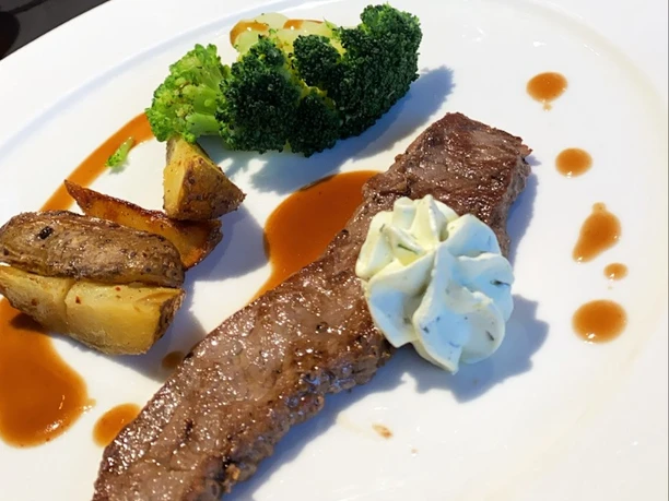 Fleischgericht Ein Stück gegrilltes Rindersteak mit Kräuterbutter, Kartoffelecken und Brokkoli auf weißem Teller.