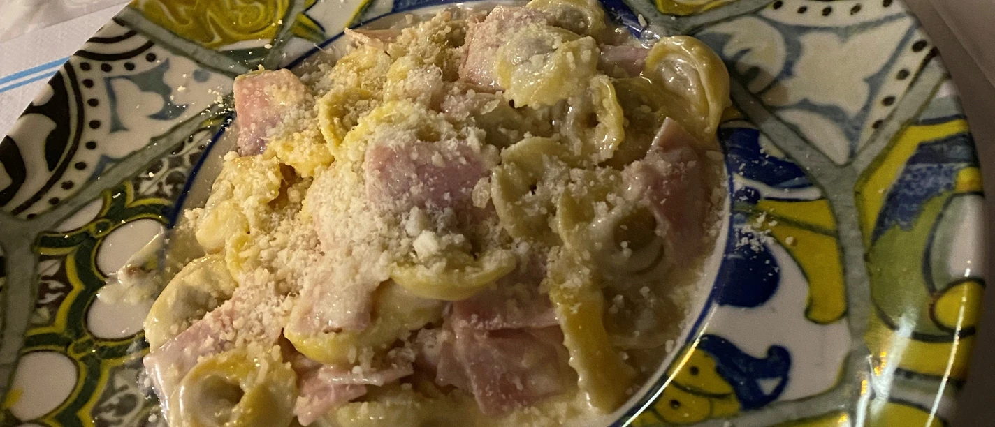Een bord gevuld met tortellini en stukjes ham, overgoten met een romige kaassaus.