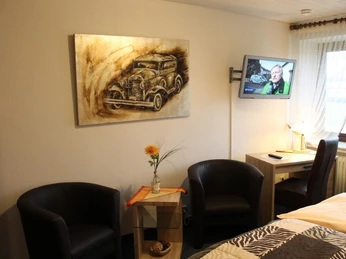 Hotel-Restaurant Lindenhof Zwei Ledersessel neben Tisch mit Blume, antikes Auto als Gemälde an Wand, Fernseher eingeschaltet.