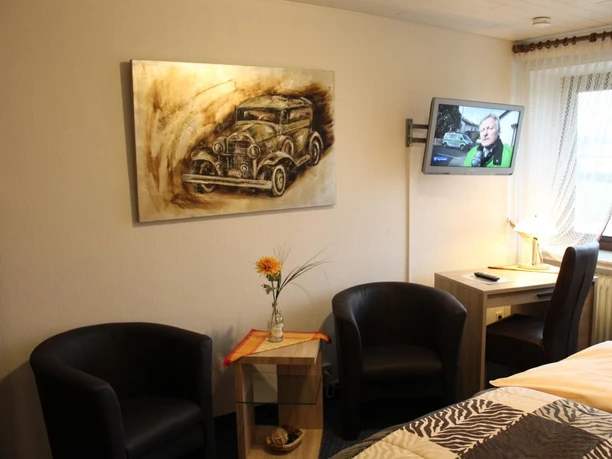 Hotel-Restaurant Lindenhof Zwei Ledersessel neben Tisch mit Blume, antikes Auto als Gemälde an Wand, Fernseher eingeschaltet.