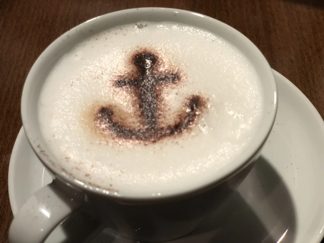 Eine weiße Kaffeetasse mit Milchschaum, darauf ein Anker aus Kakaopulver als Dekoration.A white coffee cup with milk foam and an anchor made of cocoa powder as decoration.En hvid kaffekop med mælkeskum og et anker lavet af kakaopulver som dekoration.Een witte koffiekop met melkschuim en een anker van cacaopoeder als decoratie.