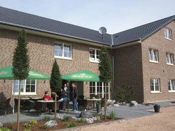 Landhotel-zum-gruenen-Kranze_4.jpg Außenansicht eines zweistöckigen Hotels aus Ziegeln mit Terrasse, Tischen und grünen Sonnenschirmen.