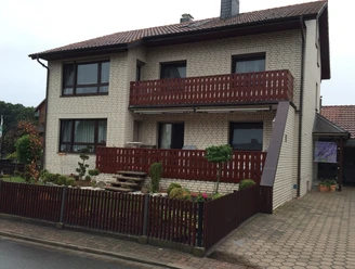 Gaestehaus-Witting_1.jpg Zweistöckiges Gästehaus mit Backsteinfassade, überdachtem Balkon und kleinem Vorgarten mit Zaun.