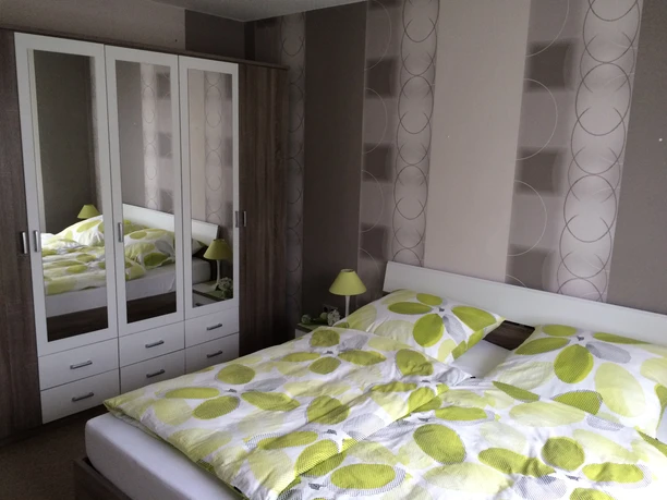 Gaestehaus-Witting_2.jpg Ein modernes Schlafzimmer mit Doppelbett, eleganter Tapete, grün-gelben Bettbezügen und Kleiderschrank.