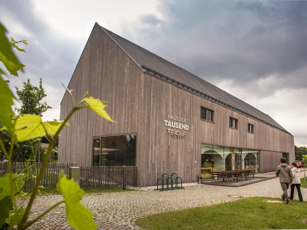 Aussenansicht Moderne Holzarchitektur mit großen Fenstern, Menschen spazieren vor einem bewölkten Himmel.