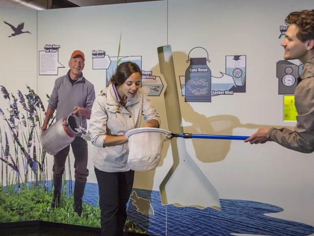 Ausstellung Zwei Personen in wetterfester Kleidung angeln in einer interaktiven Ausstellungshalle.