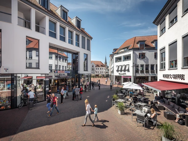 Fußgängerzone mit Geschäften und Cafés, Menschen spazieren und sitzen bei sonnigem Wetter an Tischen.