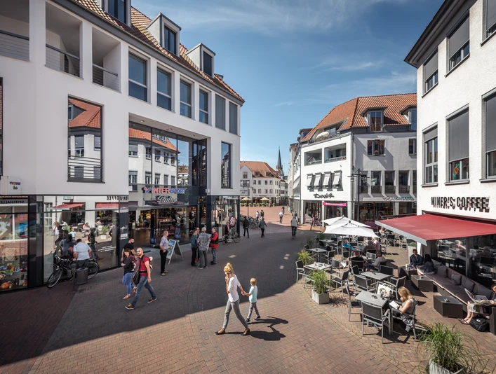 Fußgängerzone mit Geschäften und Cafés, Menschen spazieren und sitzen bei sonnigem Wetter an Tischen.