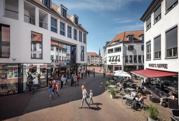 Buntes Treiben auf dem Kolbeplatz Gütersloh Fußgängerzone mit Geschäften und Cafés, Menschen spazieren und sitzen bei sonnigem Wetter an Tischen.