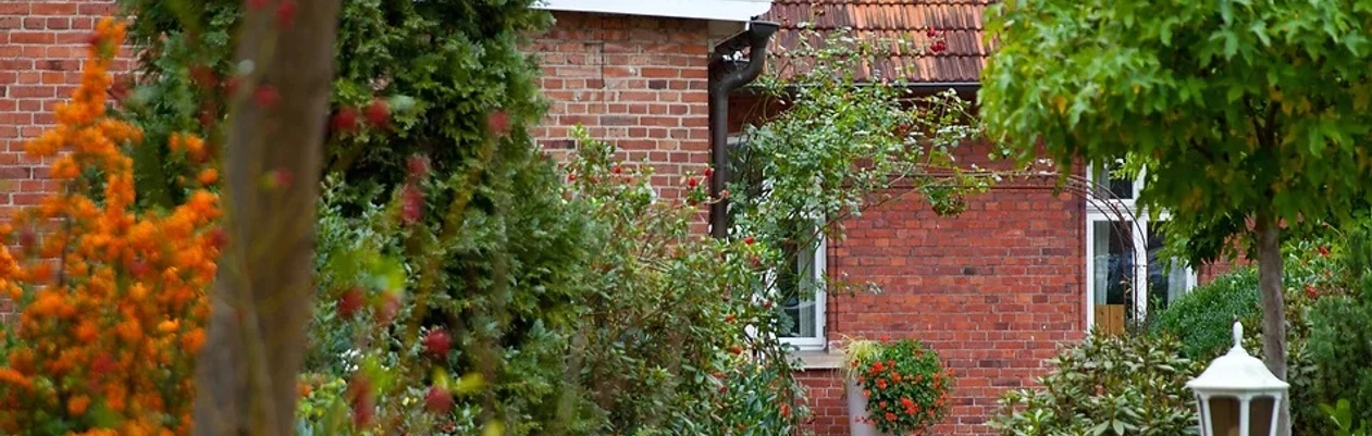 Gasthaus Rose - Hotel Alte Schule .jpg Rotes Backsteingebäude mit blühenden Pflanzen und Bäumen umgeben, ruhige Umgebung, Laterne.