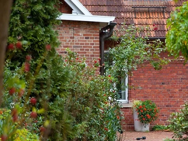 Gasthaus Rose - Hotel Alte Schule .jpg Rotes Backsteingebäude mit blühenden Pflanzen und Bäumen umgeben, ruhige Umgebung, Laterne.