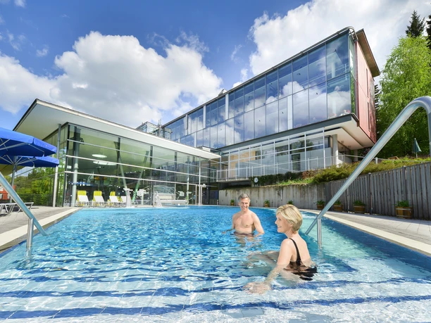 Swimming pool with glass building in the background, two people in the water under a blue sky.