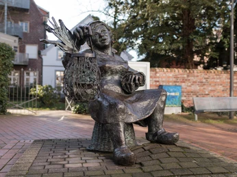 Güths Mariechen Bronzestatue einer sitzenden Frau mit einem Netz voll Fischen. Sie lächelt entspannt im Freien.