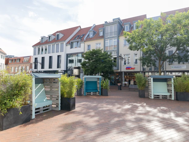 Sommerlandschaft Kolbeplatz Ein sonniger Platz mit modernen, mehrstöckigen Gebäuden, umgeben von Bäumen und bepflanzten Sitzmöglichkeiten.