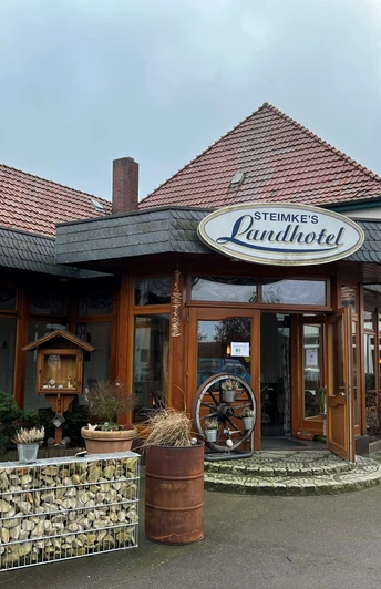 Eingang von Steimkes Landhotel mit runden Fenstern, Schindeldach und dezent begrünten Pflanzkübeln.Entrance to Steimke's country hotel with round windows, shingle roof and discreetly planted planters.Indgang til Steimkes landhotel med runde vinduer, spåntag og diskret beplantede krukker.Ingang van Steimke's plattelandshotel met ronde ramen, leien dak en discreet beplante plantenbakken.