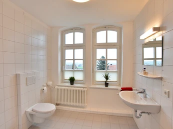 Alte Lübber Volksschule Ein modernes, weiß gefliestes Badezimmer mit zwei Fenstern, Waschbecken, Spiegel und WC.