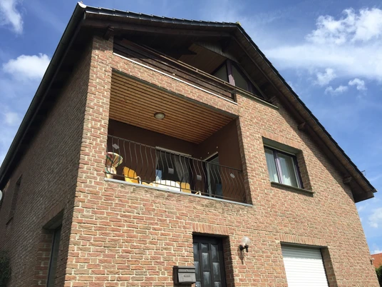 Ferienwohnungen-Dahme2.JPG Zweistöckiges Backsteinhaus mit Balkon, Dachfenstern und geschwungenem Treppengeländer unter blauem Himmel.