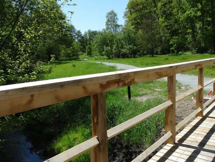 De-luhpadd-1-wanderweg-foto_ccbysa_foto©gemeinde-bispingen.jpg Holzbrücke mit Geländer überquert sanften Bach, umgeben von grünem Wald im Sonnenlicht.