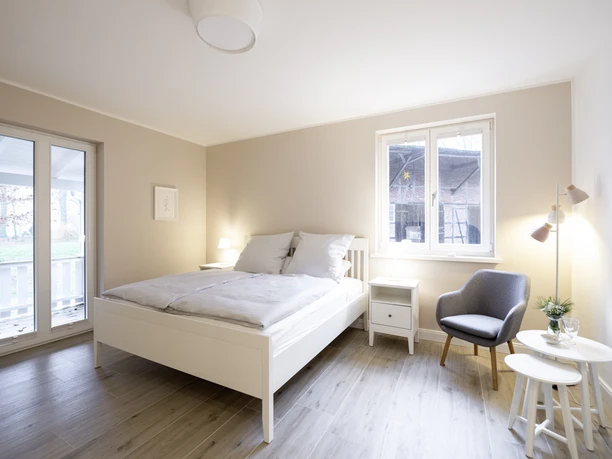 Doppelzimmer mit weißem Bett Helles Doppelzimmer mit weißem Bett, grauem Sessel, Holzboden und verglaster Terrassentür.