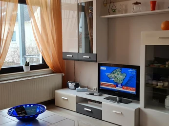 Gemütliches Wohnzimmer Wohnzimmer mit großen Fenstern, hellen Vorhängen, modernem TV-Möbel und Dekorationen in Erdtönen.