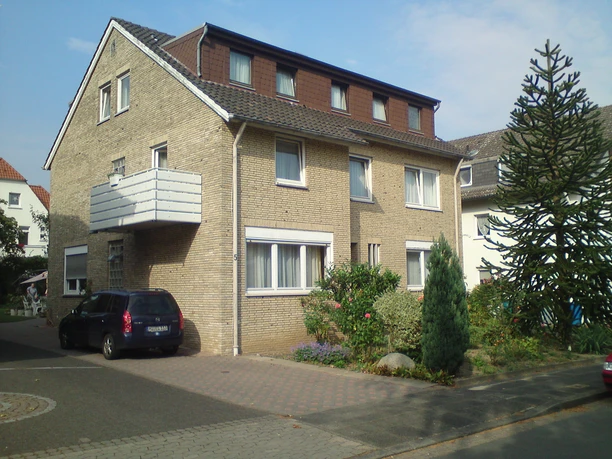 Außenansicht Mehrfamilienhaus aus gelben Backsteinen mit drei Etagen und einem Balkon, umgeben von kleinen Gärten.