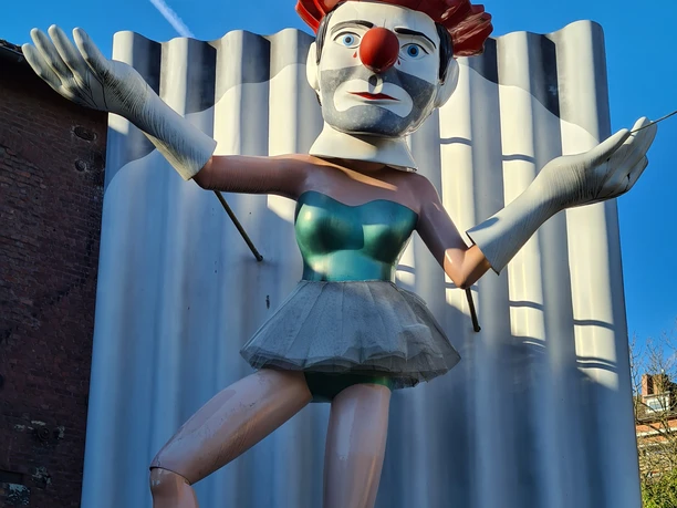 Clownsfigur mit rotem Hut, blauer Bluse und erhobenen Armen.