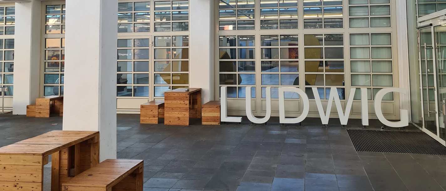 Innenhof des Ludwig Forums mit moderner Architektur mit hohen, weißen Säulen und großen Fenstern. Große Buchstaben, die das Wort "LUDWIG" formen, stehen auf dem Boden.