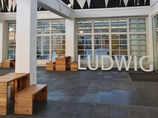 Innenhof des Ludwig Forums mit moderner Architektur mit hohen, weißen Säulen und großen Fenstern. Große Buchstaben, die das Wort "LUDWIG" formen, stehen auf dem Boden.