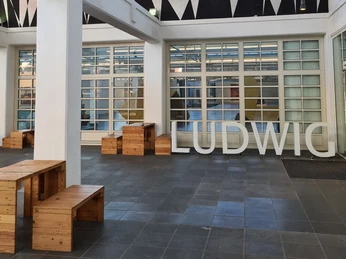 Ludwig Forum Innenhof des Ludwig Forums mit moderner Architektur mit hohen, weißen Säulen und großen Fenstern. Große Buchstaben, die das Wort "LUDWIG" formen, stehen auf dem Boden.