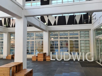 Ludwig Forum Innenhof des Ludwig Forums mit moderner Architektur mit hohen, weißen Säulen und großen Fenstern. Große Buchstaben, die das Wort "LUDWIG" formen, stehen auf dem Boden.