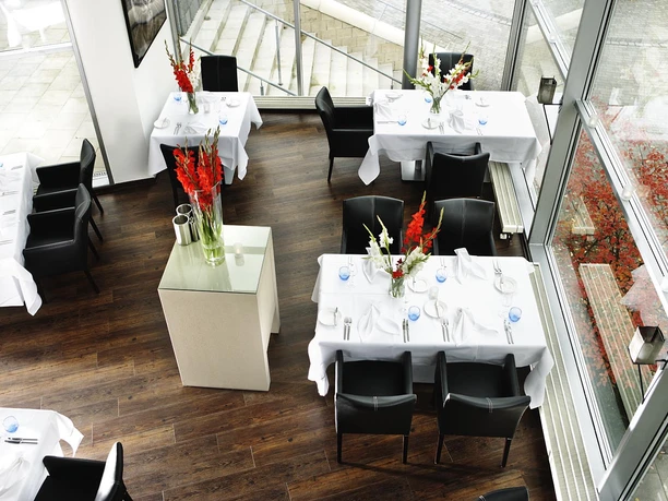 _U3Q0126b.jpg Zu sehen ist die stillvolle Restauranteinrichtung aus einer erhöhten Perspektive. Die weißen, elegant gedeckten Tische mit schwarzen Stühlen stehen in einem modernen lichtdurchfluteten Raum mit großen Glasfenstern, die den Blick auf eine Außenterrasse ermöglichen.
