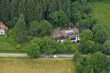 Gesundheitshaus-am-Wiehengebirge.jpg Luftaufnahme eines Hauses am Waldrand mit Solarpanels auf dem Dach, umgeben von Bäumen und Feldern.