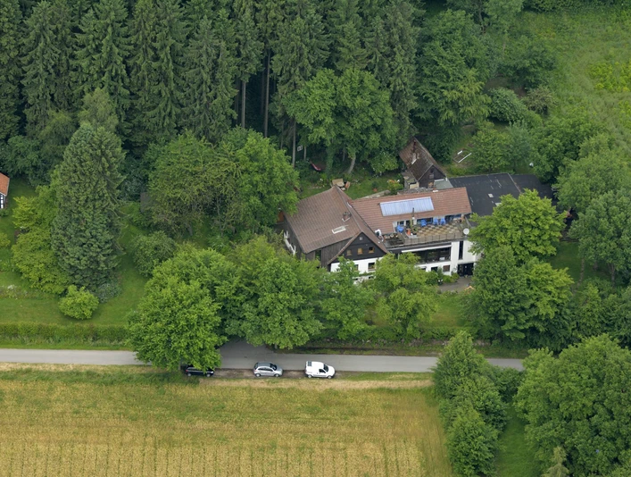 Gesundheitshaus-am-Wiehengebirge.jpg Luftaufnahme eines Hauses am Waldrand mit Solarpanels auf dem Dach, umgeben von Bäumen und Feldern.